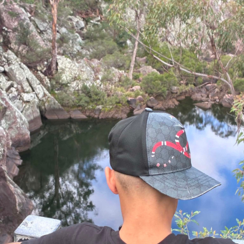 A person wearing an Indigenous-designed snapback cap looks out over a still, rock-lined waterhole surrounded by native bushland and reflected gum trees.